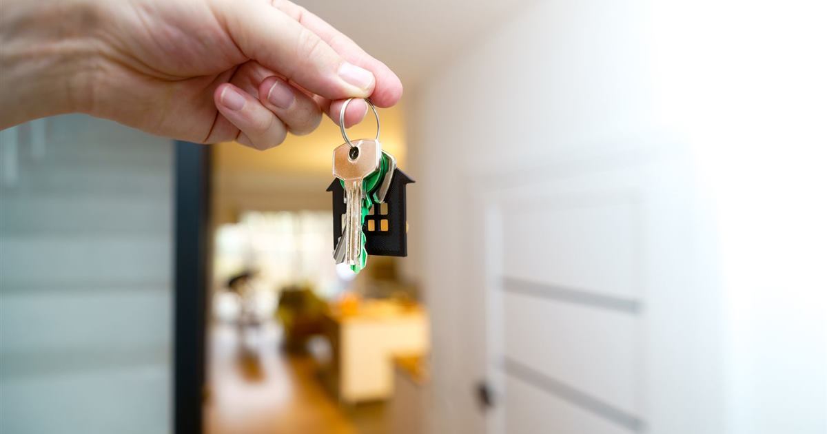 A hand holding up house keys on a house-shaped key ring in the doorway of an empty flat