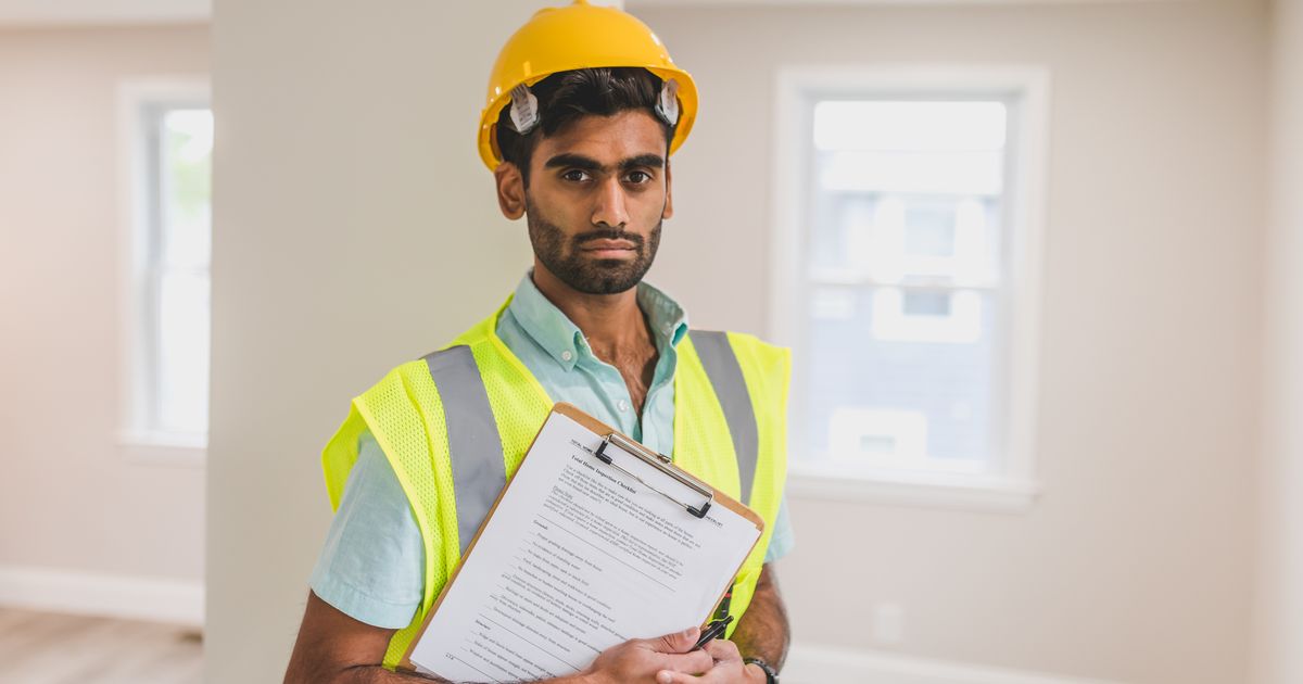 A man in a yellow hard hat and safety vest holding a clipboard indoors