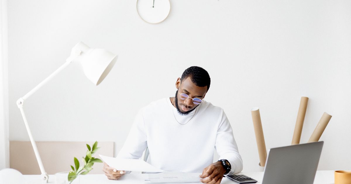 A man in glasses reading papers at a white desk with a laptop, calculator and desk lamp