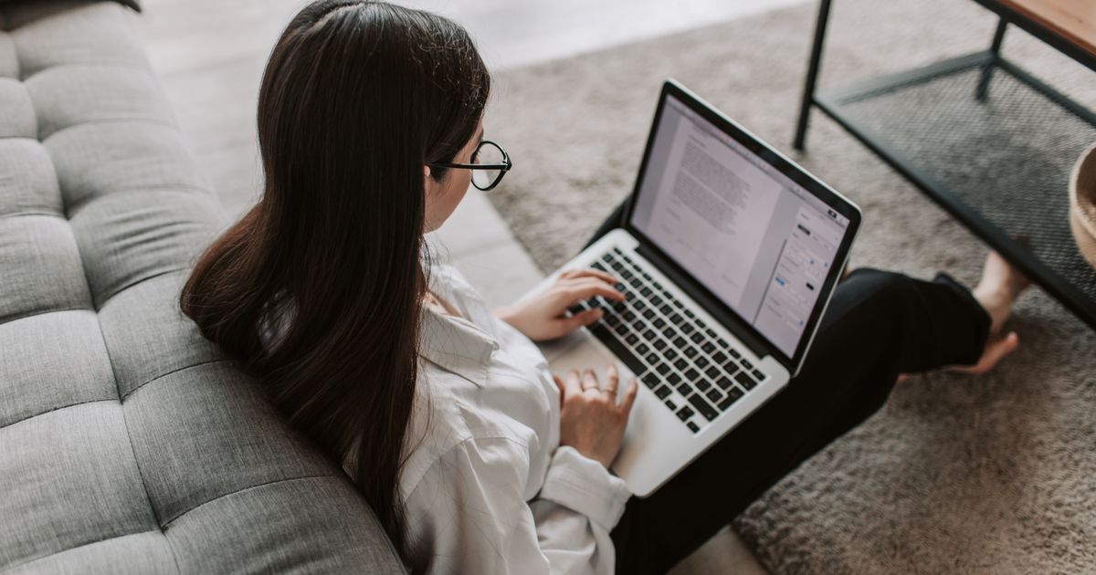 A woman in glasses sitting on the floor against a grey couch typing on a laptop