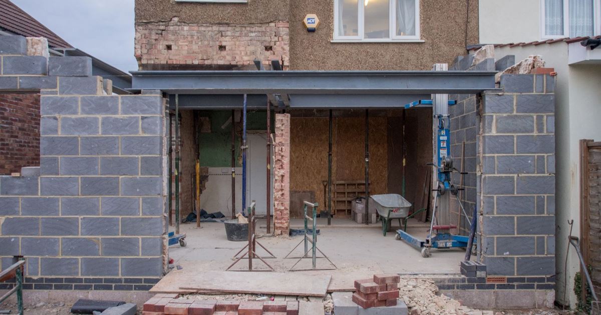 A brick house with a new block extension going up at the back, steel beams and a wheelbarrow on the slab