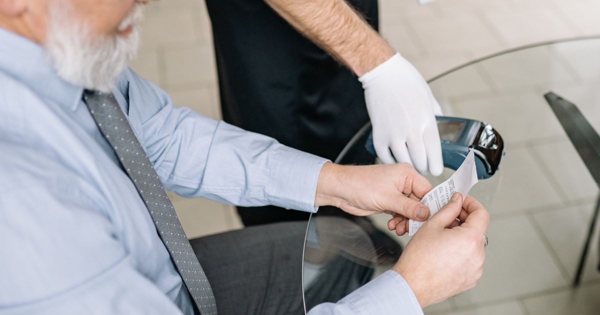 An older man pays by card at a restaurant table while a gloved server holds the card machine