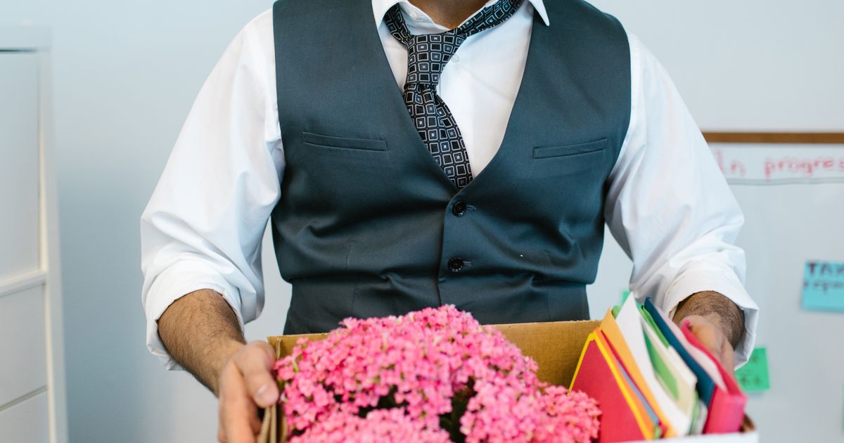 A man in a waistcoat carrying a cardboard box of folders and a pink pot plant out of an office