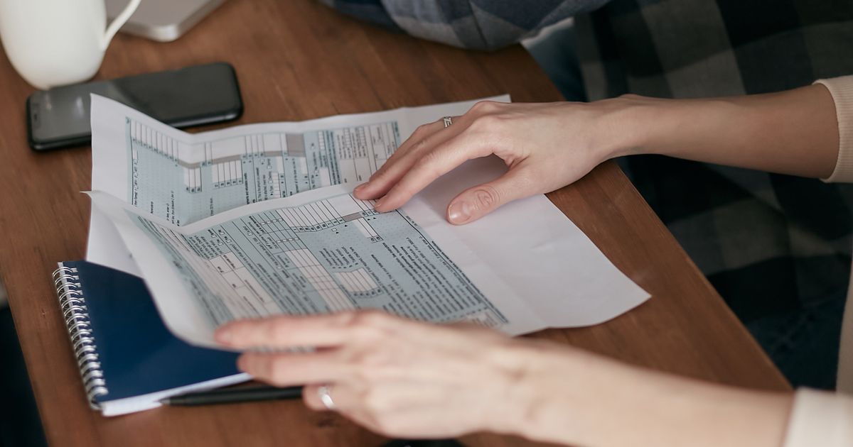 Hands holding tax forms on a wooden table next to a calculator and a laptop