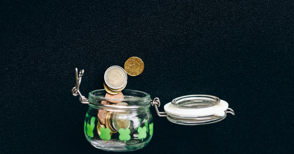 Coins dropping into an open glass jar decorated with green clovers on a black background
