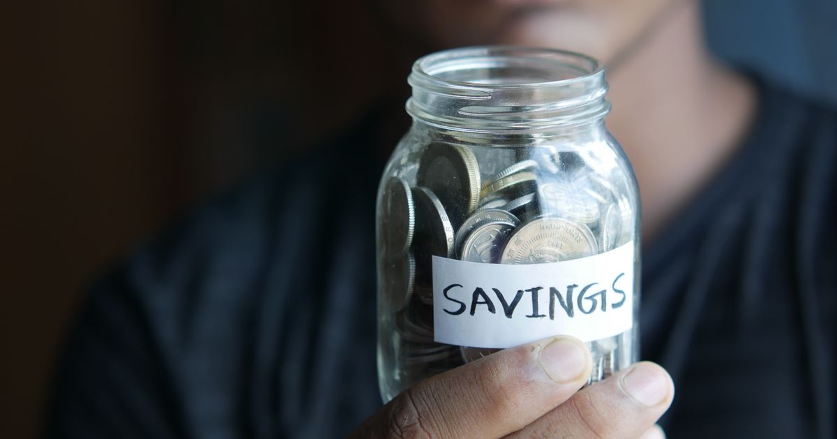 A hand holding a glass jar of coins with a paper label that reads Savings