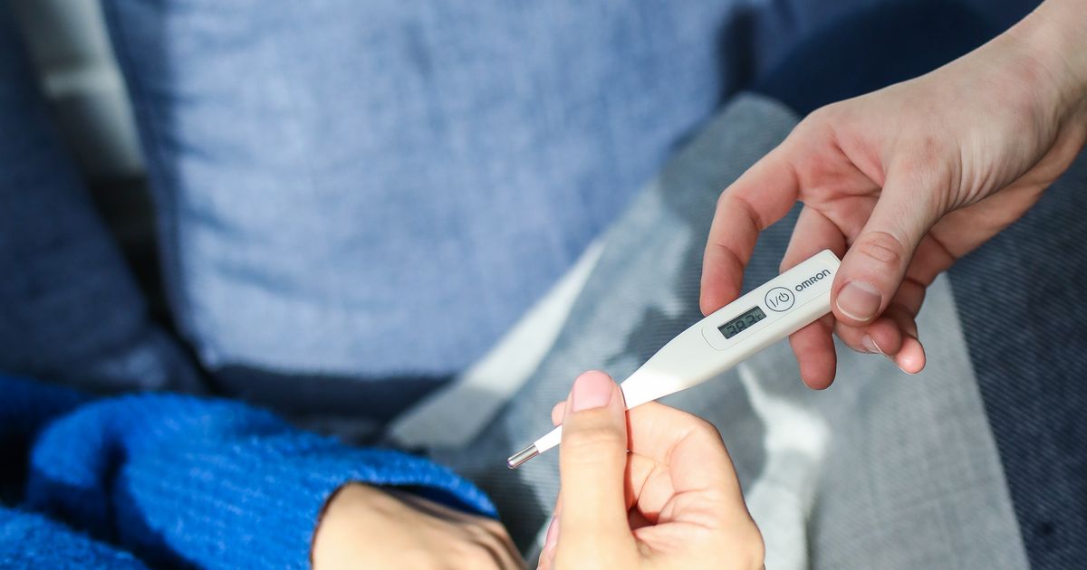 A woman wrapped in a blanket on a couch checking her temperature with a thermometer