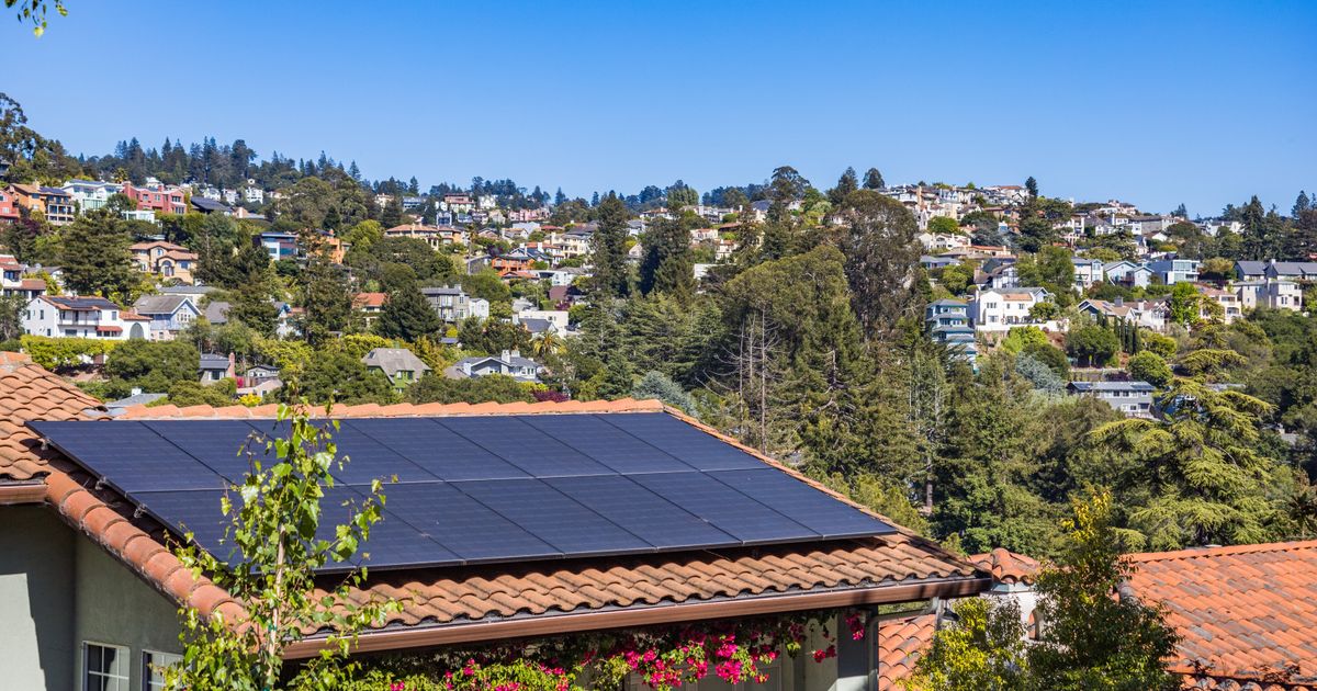 Black solar panels on a terracotta tiled roof with a hillside of houses behind it under a blue sky